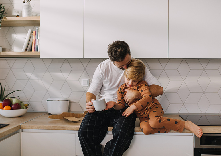 Dad cuddling with toddler son as they sit on top of the kitchen counter
